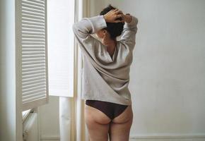 Self loving attractive woman plus size in knitted cardigan and underwear from back near window at light room, body love photo