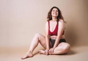 Self loving young woman plus size in underwear sitting on beige background, body love photo