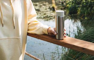 Brunette woman in yellow hoodie with thermo mug relaxing near lake in early morning photo