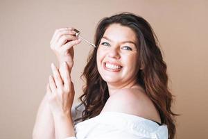 Beauty portrait of body positive woman with dark hair with pipette with organic oil on clean fresh skin face and hands on beige background photo