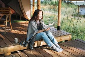 Young brunette woman in poncho reading book and relaxing in glamping in the woods photo