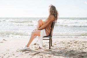 joven hermosa mujer despreocupada con el pelo largo en suéter sentado en una silla en la playa del mar foto