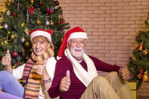 pareja caucásica mayor celebrando la navidad juntos en felicidad y emoción en casa con sombrero rojo de santa y árbol de navidad foto