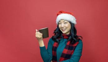 Pretty smiling asian woman in warm christmas sweater and santa hat is having hot chocolate on red background for season celebration concept photo
