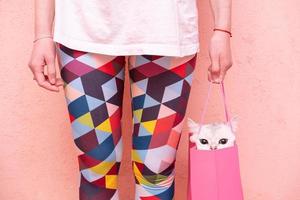 A funny cat sits in a gift bag held by a woman in multi-colored tights, on a pink background. Lifestyle. photo