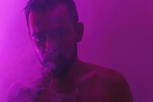 Handsome young man with wet skin in ultraviolet light smoking vape photo