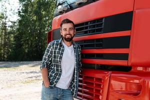 joven conductor de camión masculino sonriente al lado de su camión de carga rojo foto