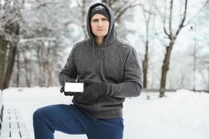 Young man athlete showing smartphone with a blank screen during winter workout photo