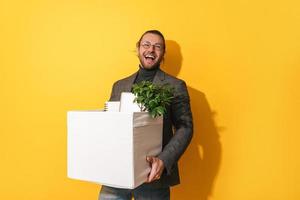 Happy man holding box with personal items after job promotion against yellow background photo