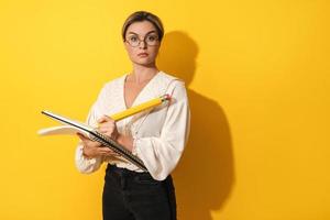 mujer seria con gafas sosteniendo un lápiz grande y un cuaderno sobre fondo amarillo foto