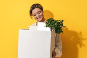 Happy woman holding box with personal items after job promotion against yellow background photo