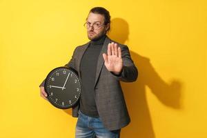 Serious man holding clock and showing stop gesture on yellow background photo