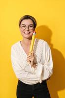 Cheerful woman wearing glasses holding big pencil on yellow background photo