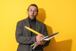 Funny man wearing glasses holding big pencil and notebook on yellow background photo