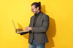 Happy bearded man wearing eyeglasses is using laptop computer on yellow background photo
