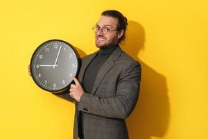 Anxious man wearing glasses holding big clock on yellow background photo