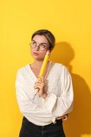 Thoughtful woman wearing glasses holding big pencil on yellow background photo