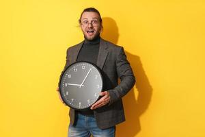 Surprised man wearing glasses holding big clock on yellow background photo