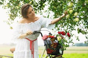 Woman on the bicycle is picking fresh apples from the tree in the village garden photo