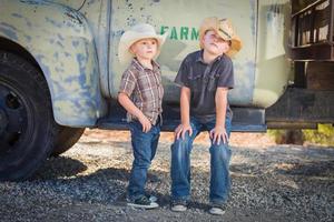 Two Young Boys Wearing Cowboy Hats Leaning Against an Antique Truck in a Rustic Country Setting. photo