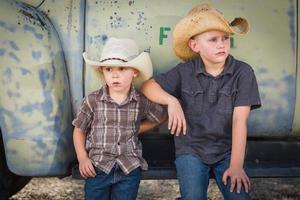 Two Young Boys Wearing Cowboy Hats Leaning Against an Antique Truck in a Rustic Country Setting. photo