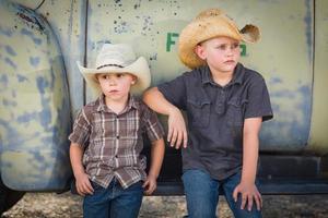 Two Young Boys Wearing Cowboy Hats Leaning Against an Antique Truck in a Rustic Country Setting. photo