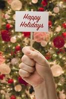 mano que sostiene la tarjeta de felices fiestas frente al árbol de navidad decorado. foto