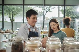 Asian male shopkeeper describes natural organic products to White woman customer in refill store, zero-waste and plastic-free grocery, environment-friendly, sustainable lifestyles in reusable shop. photo