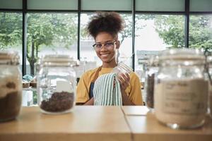 A young Black female shopkeeper cleans glass jars of natural organic products in reusable containers at a refill store, zero waste, a plastic-free grocery shop, and an eco-friendly retail business. photo