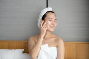 Happy young Asian woman applying face lotions while wearing a towel and touching her face. Daily makeup and skincare photo