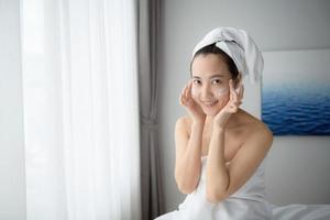 Happy young Asian woman applying face lotions while wearing a towel and touching her face. Daily makeup and skincare photo