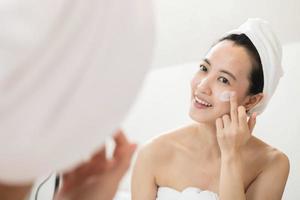 Happy young Asian woman applying face lotions while wearing a towel and touching her face in bathroom photo