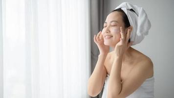 Happy young Asian woman applying face lotions while wearing a towel and touching her face. Daily makeup and skincare photo
