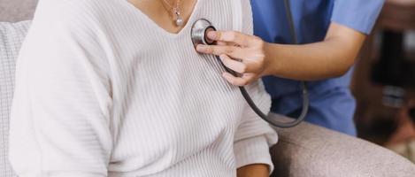 Homecare nursing service and elderly people cardiology healthcare. Close up of young hispanic female doctor nurse check mature caucasian man patient heartbeat using stethoscope during visit photo