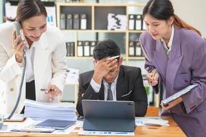 A team of three Asian businessmen is working inside an office where businessmen in black suits are being ordered by their supervisors and bosses a lot causing stress at work. burnout concept photo