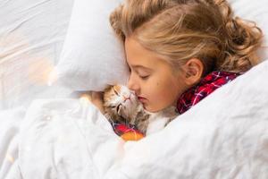 a cute little girl is sleeping sweetly at home in a rabbit with a kitten. White cotton bed linen. Christmas holidays. Children and pets at home photo