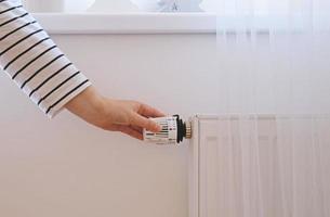 Woman regulates temperature at home with the heating thermostat to save energy, close-up with hand. Temperature control valve on radiator. White heater on wall with curtain in modern apartment. photo