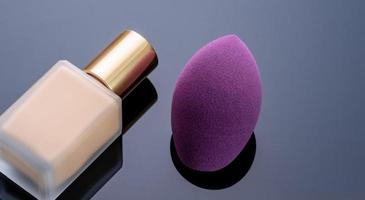 Purple makeup sponge on dark background. Soft makeup cosmetic sponge. Flat-ended makeup sponge with blur makeup foundation bottle. Liquid beige make-up foundation and beauty blender. Cosmetic product. photo