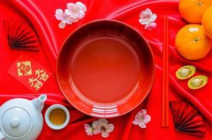 plato rojo con palillos sobre fondo de tela satinada roja con juego de té, lingotes, naranjas y paquete de sobre rojo o palabra ang bao significa riqueza, suerte para el concepto de cena de año nuevo chino. foto