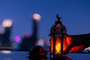 enfoque selectivo de la linterna que tiene el símbolo de la luna en la parte superior y un pequeño plato de dátiles con fondo de ciudad para la fiesta musulmana del mes sagrado del ramadán kareem. foto