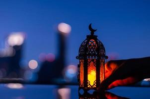 enfoque selectivo de la linterna que tiene el símbolo de la luna en la parte superior sosteniendo a mano con el fondo de la ciudad para la fiesta musulmana del mes sagrado del ramadán kareem. foto