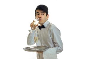 un joven camarero con una camisa blanca con una mariposa sostiene una bandeja y bebe champán de una copa foto
