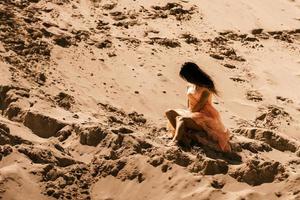 pretty brunette girl in pink dress sits on sand photo