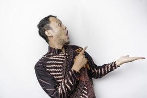 Shocked Asian man wearing batik shirt pointing at the copy space beside him, isolated by white background photo