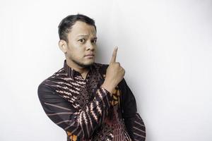 Serious Asian man wearing batik shirt pointing at the copy space on top of him, isolated by white background photo