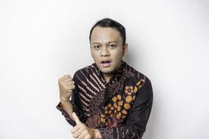 Shocked Asian man wearing batik shirt pointing at the copy space beside him, isolated by white background photo