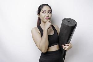 una mujer asiática deportiva y pensativa que usa ropa deportiva sostiene una alfombra de yoga en un fondo blanco aislado. foto