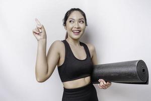 Excited sporty Asian woman wearing sportswear pointing at the copy space on top of her while carrying yoga mat, isolated by white background photo