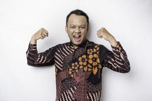 Excited Asian man wearing batik shirt showing strong gesture by lifting his arms and muscles smiling proudly photo