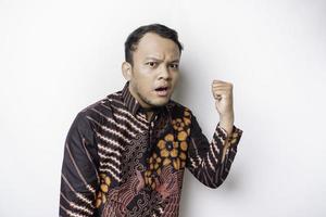 Shocked Asian man wearing batik shirt pointing at the copy space beside him, isolated by white background photo